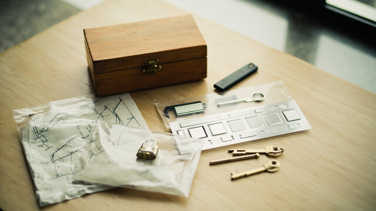 A closed wooden box with a brass clasp on a pale oak table, beside a hand-drawn tree diagram sealed in a clear bag with a small padlock, a steel backup plate, a USB stick and three brass keys.